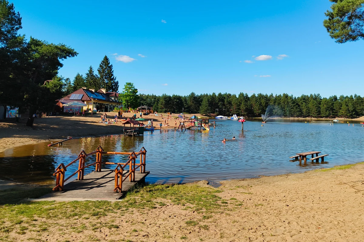 Plaża otoczona lasem z kąpieliskiem, pomostem i grupą osób korzystających z uroków letniego dnia; na drugim planie budynek rekreacyjny oraz fontanna w wodzie.