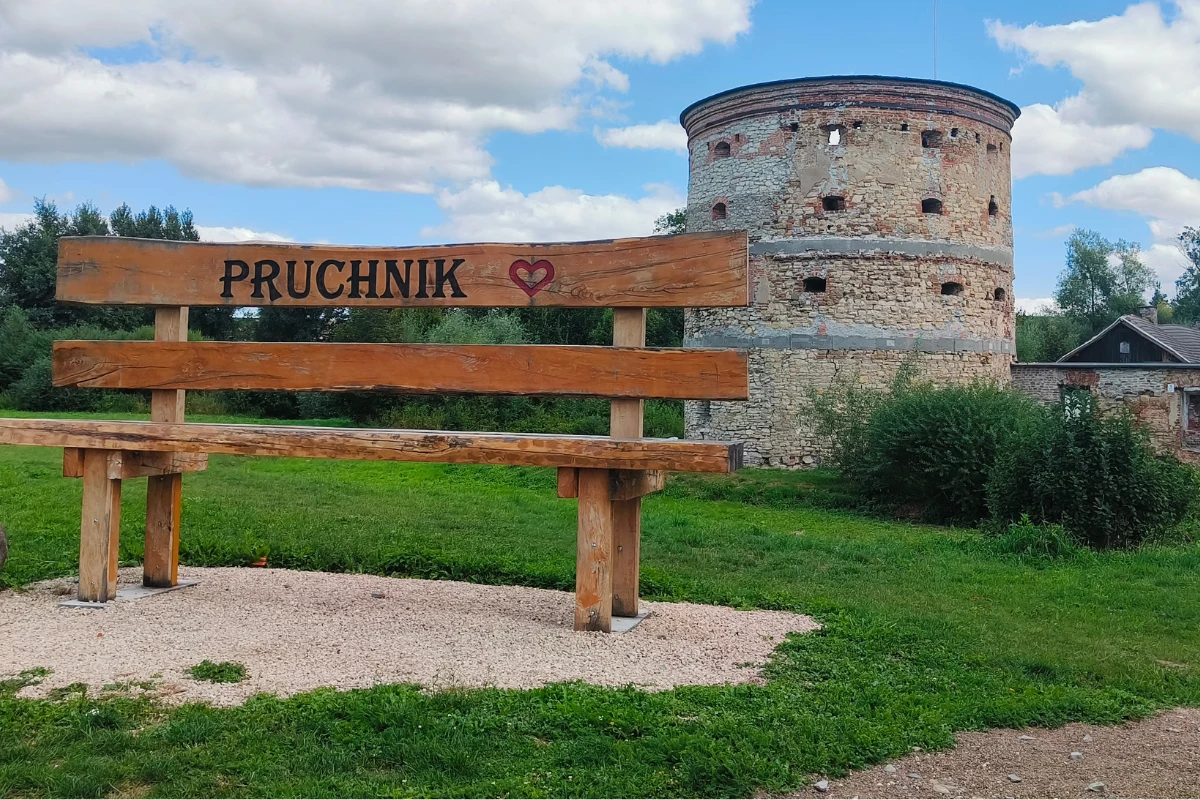 Drewniana ławka z napisem „Pruchnik” i sercem ustawiona przed zabytkową, okrągłą wieżą z kamienia na tle zieleni i nieba z chmurami.