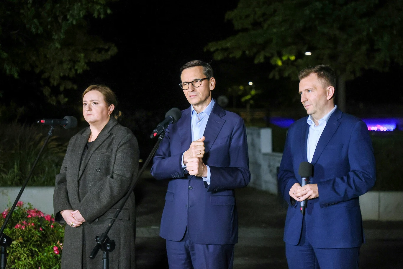 Olga Semeniuk, Mateusz Morawiecki i Łukasz Schreiber podczas wieczornej konferencji prasowej Rozwoju Plus w Warszawie. Morawiecki stoi przy mikrofonie między politykami.
