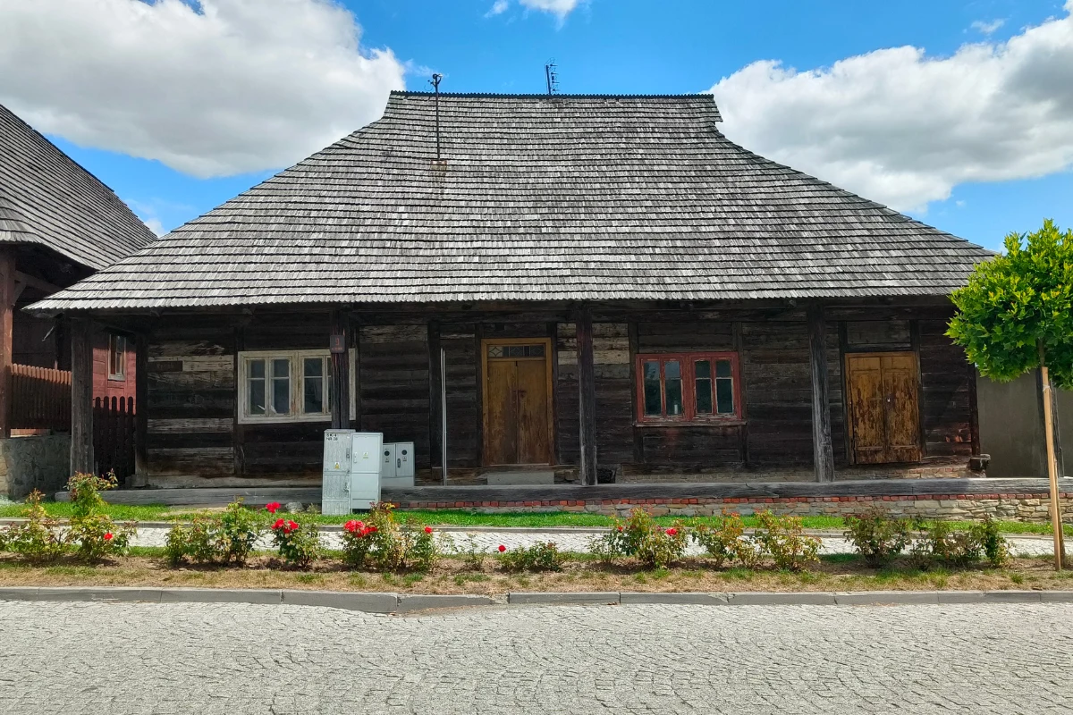 Drewniany dom z ciemnymi belkami, dwuspadowym gontowym dachem i trzema wejściami, przed budynkiem niska rabata z różami oraz brukowany chodnik.