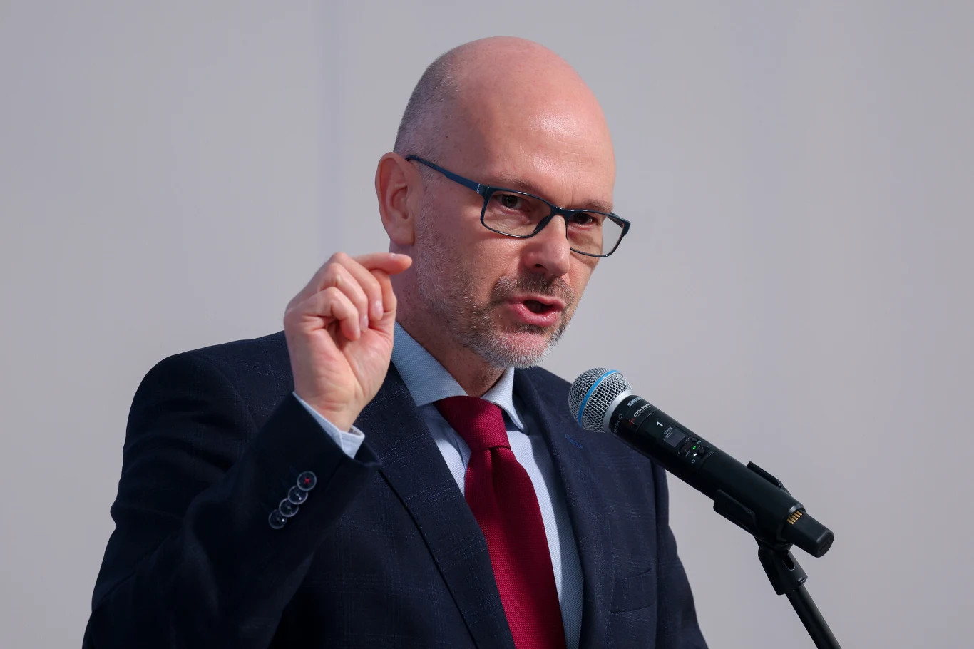 Mężczyzna w garniturze i okularach wygłaszający przemówienie do mikrofonu, gestykulujący uniesioną dłonią.