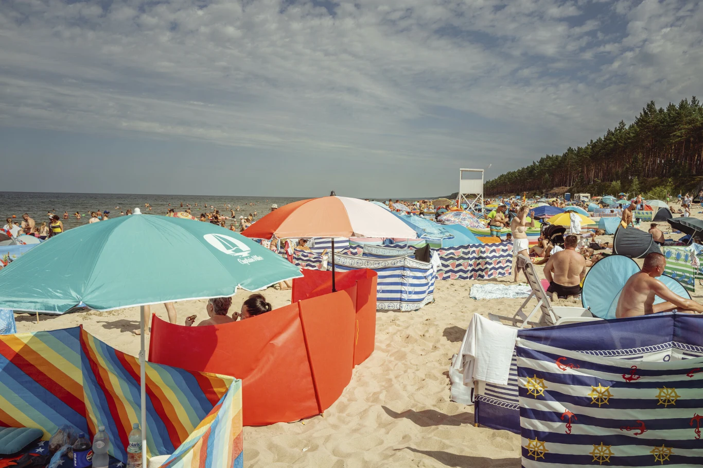 Ile wziąć urlopu, żeby dostać wczasy pod gruszą? Plaża z wieloma osobami, kolorowe parawany i parasole, las sosnowy i wieża ratownika w tle.