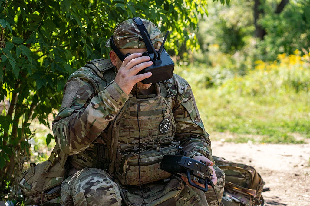 Ukraiński operator dronów Żołnierz w mundurze maskującym siedzi pod drzewem, trzymając w jednej ręce kontroler drona, a drugą poprawiając gogle do obsługi zdalnej technologii.