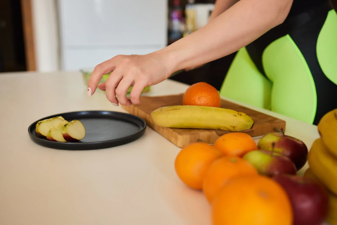 Owoce w kuchni mogą przyciągać muszki owocówki. Ręka sięga po plasterek jabłka na talerzu wśród owoców, w tym banana i pomarańczy, na kuchennym blacie.
