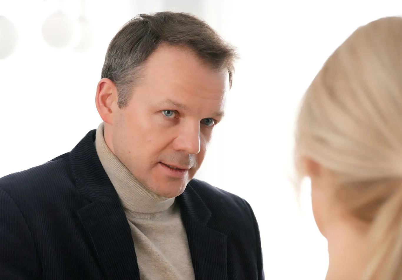 Mężczyzna w ciemnej marynarce i jasnym golfie patrzy poważnie na blondynkę stojącą naprzeciwko.
