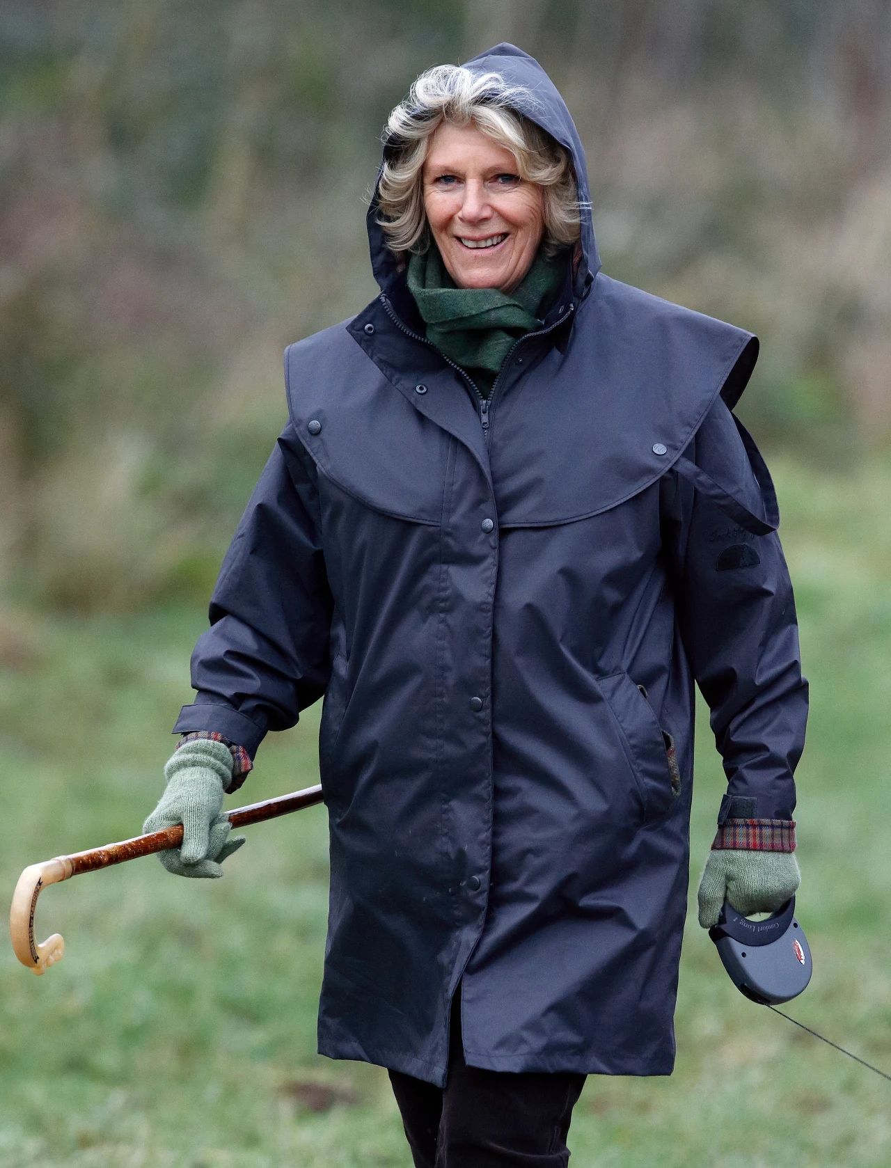 Królowa Camilla rzadko widywana jest w prywatnych sytuacjach, gdzie jej strój jest zdecydowanie mniej formalny Uśmiechnięta kobieta w ciemnym płaszczu przeciwdeszczowym z kapturem, trzymająca laskę pasterską oraz smycz automatyczną, ubrana w zielone rękawiczki i szalik, spaceruje po trawie.