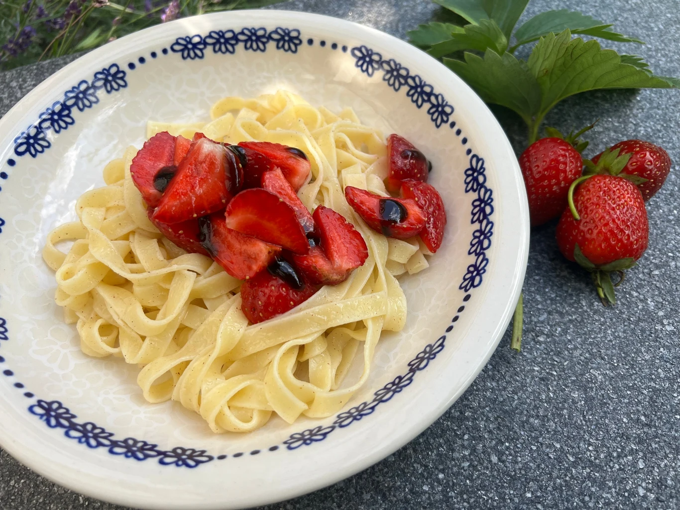 Makaron tagliatelle z truskawkami pokrojonymi na kawałki i polewą balsamiczną, podany na ozdobnym talerzu; obok świeże truskawki i zielone liście.