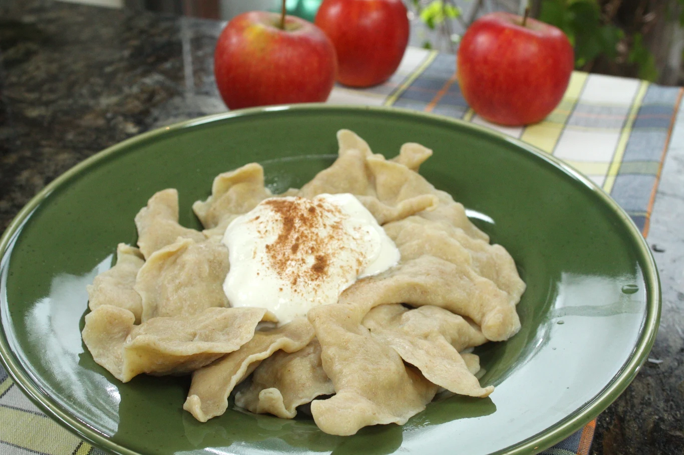 Pierogi podane na zielonym talerzu, udekorowane śmietaną posypaną cynamonem, w tle dwie czerwone jabłka.