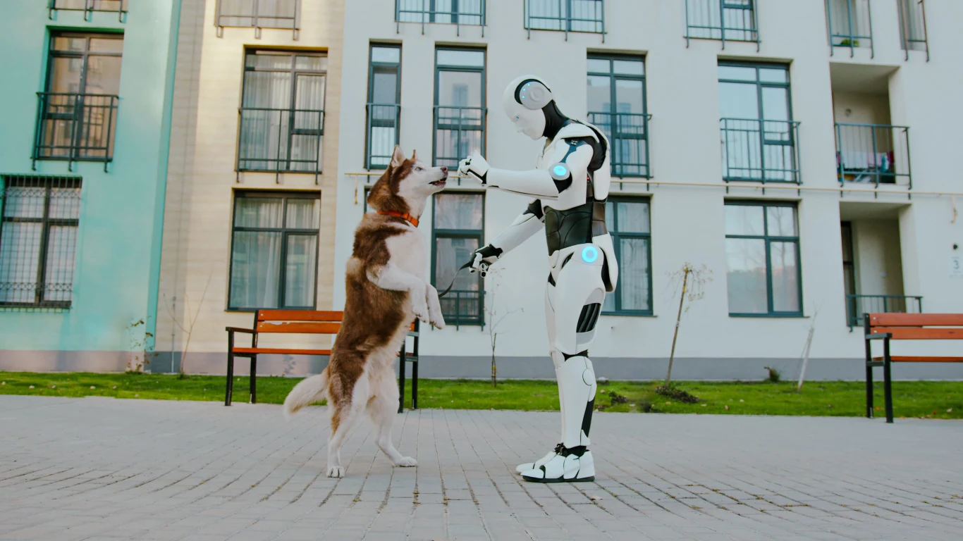 Robot w białym pancerzu trzyma za łapy dużego psa stojącego na tylnych nogach na chodniku przed budynkiem mieszkalnym.