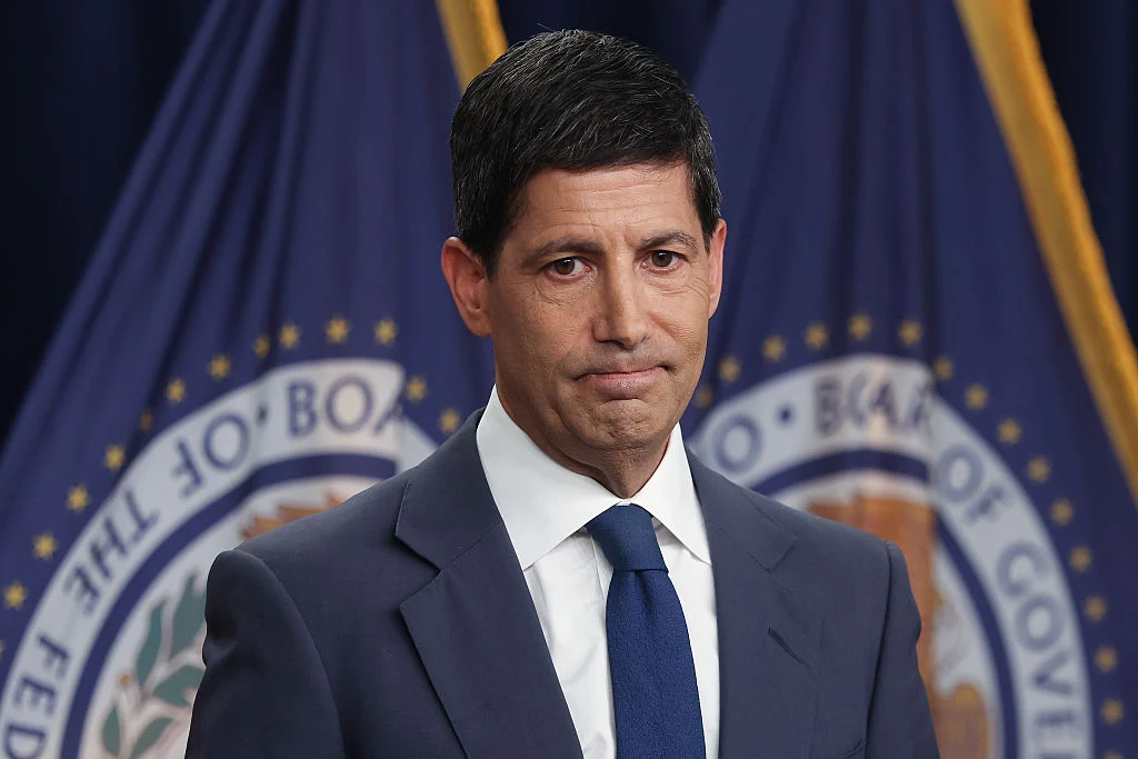 Prezes Rezerwy Federalnej Kevin Warsh Mężczyzna w garniturze i krawacie stoi przed fioletowymi flagami z oficjalnymi emblematami.