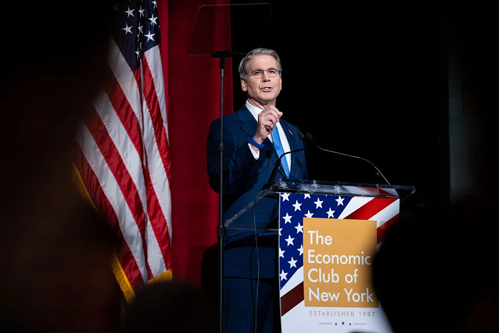 Mężczyzna w garniturze przemawia przy mównicy z logo The Economic Club of New York, na tle flagi Stanów Zjednoczonych.