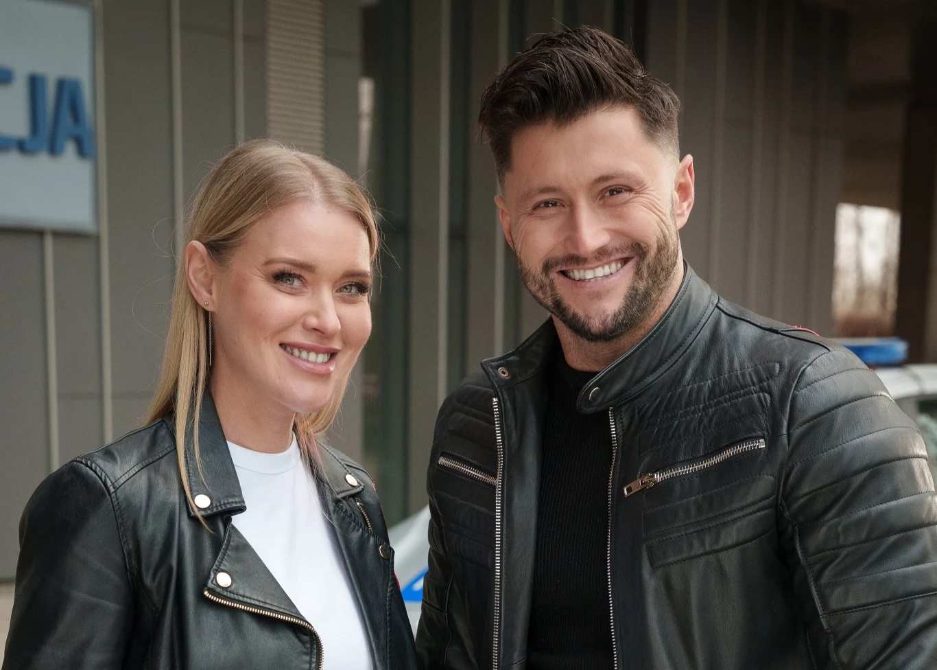 Ewelina Ruckgaber i Piotr Mróz Dwie uśmiechnięte osoby w ciemnych skórzanych kurtkach stojące obok siebie na zewnątrz budynku, w tle widoczny napis