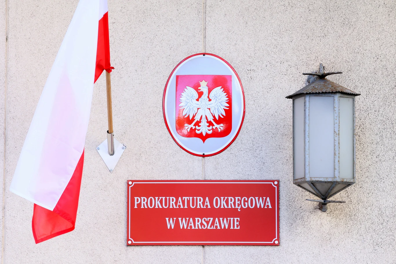 Tablica z napisem „Prokuratura Okręgowa w Warszawie”, biało-czerwona flaga Polski, godło państwowe oraz lampa ścienna.