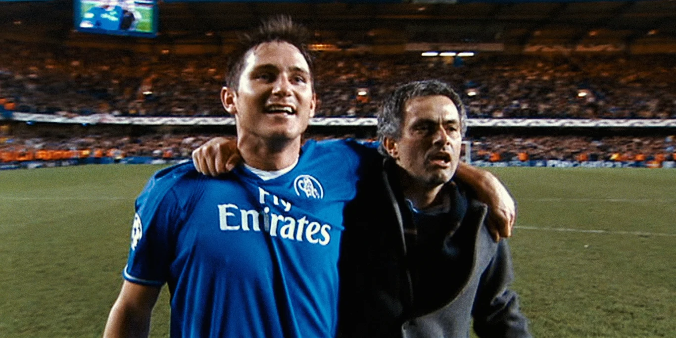 Frank Lampard i José Mourinho Piłkarz w niebieskiej koszulce obejmuje mężczyznę w ciemnej marynarce na stadionie, otoczeni przez rozświetlone trybuny pełne kibiców.