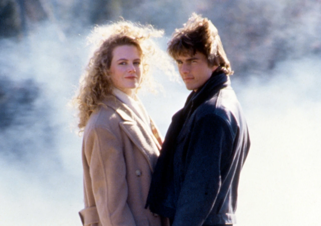 Nicole Kidman i Tom Cruise na planie filmu "Days of Thunder", rok 1990 Dwie osoby stoją obok siebie na zewnątrz, skierowane w stronę aparatu; kobieta z kręconymi, jasnymi włosami oraz mężczyzna z ciemniejszymi, rozwianymi włosami, oboje w jesiennych płaszczach, na tle jasnej, zamglonej scenerii.