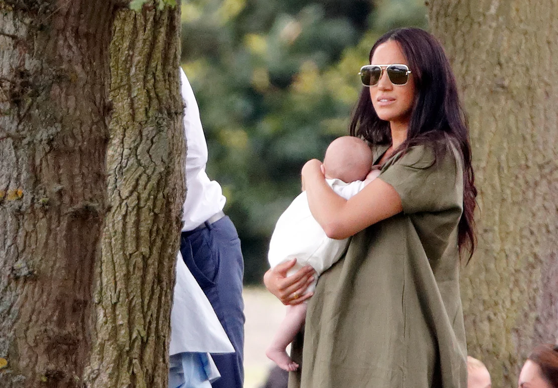 Meghan i Harry zaskoczyli wyborem imienia Kobieta w okularach przeciwsłonecznych przytula niemowlę ubrane na biało, stojąc wśród drzew.