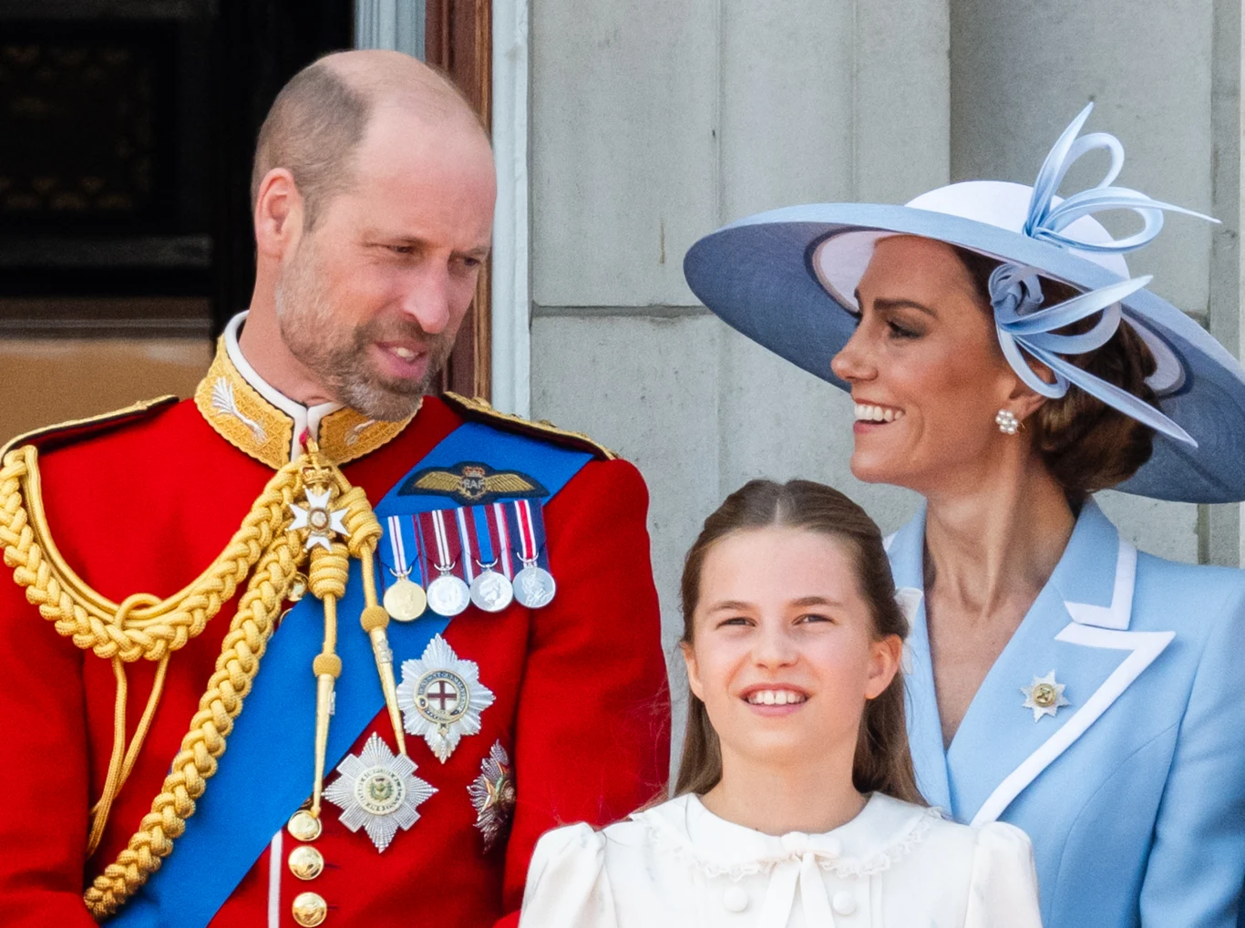 William i Kate uczcili królową w tradycyjny sposób Mężczyzna w czerwonym, paradnym mundurze z orderami stoi obok uśmiechniętej kobiety w jasnoniebieskim, eleganckim stroju i kapeluszu oraz młodej dziewczynki w białej sukience, wszyscy pozują razem na tle jasnej ściany.