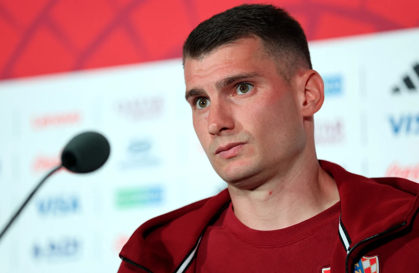 Mężczyzna w czerwonym dresie sportowym stoi przed mikrofonem na tle ściany z logotypami sponsorów.