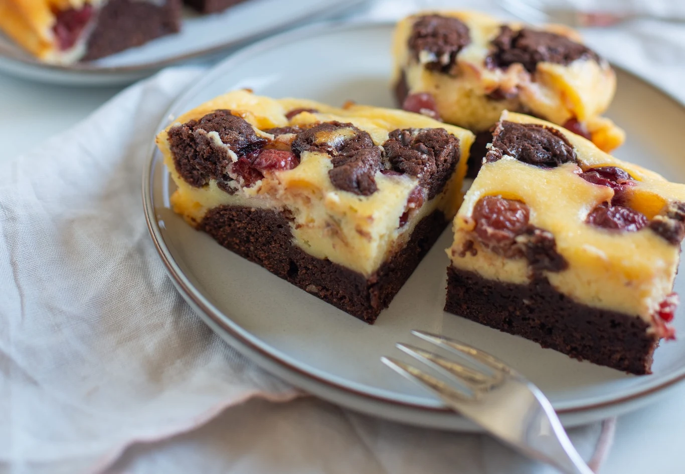 Ten deser zachwyci nie tylko wielbicieli serników Dwa kawałki sernikobrownie z wiśniami, ułożone na jasnoszarym talerzu z widelcem.