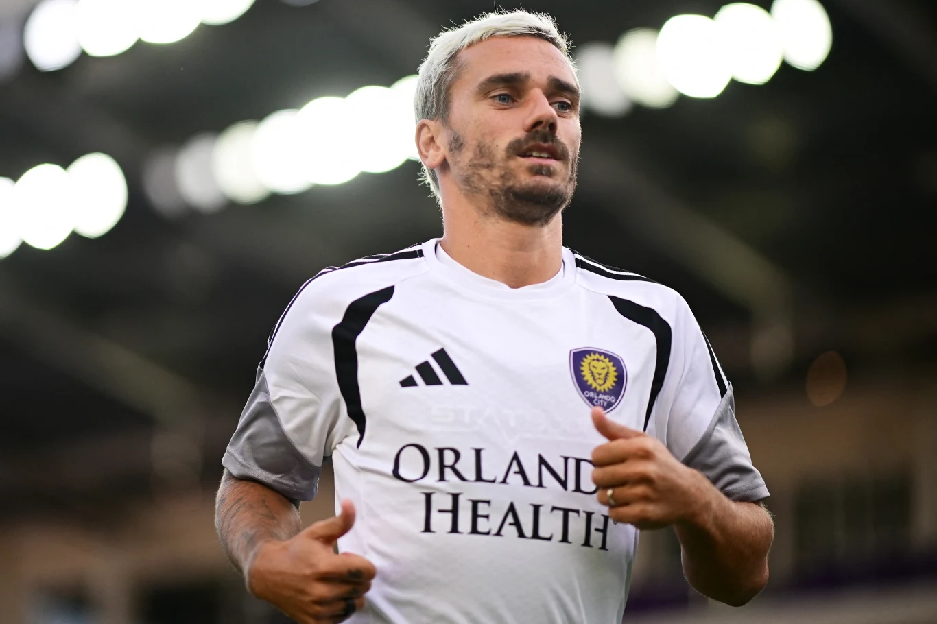 Piłkarz w białej koszulce z logo Orlando Health oraz herbem klubu Orlando City, biegnący na stadionie sportowym.