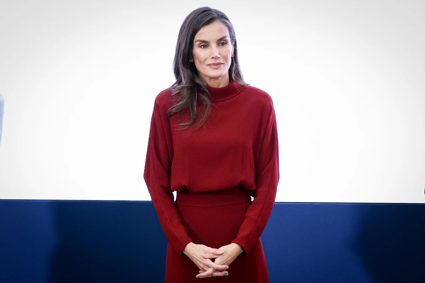 Swetry jesienią mogą wnieść odrobinę koloru. Królowa Letizia stawia na głęboką czerwień Kobieta o długich, ciemnych włosach ubrana w elegancką, bordową sukienkę stoi na tle biało-niebieskiej ściany, spoglądając lekko na bok z delikatnym uśmiechem na twarzy.