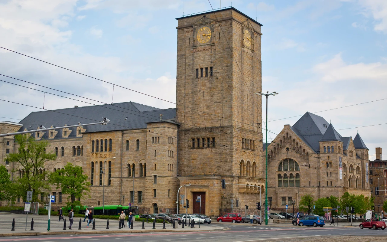 Zamek Cesarski w Poznaniu Gmach Zamku Cesarskiego w Poznaniu, z wysoką wieżą zegarową oraz detalami architektonicznymi, widoczna ulica z samochodami.