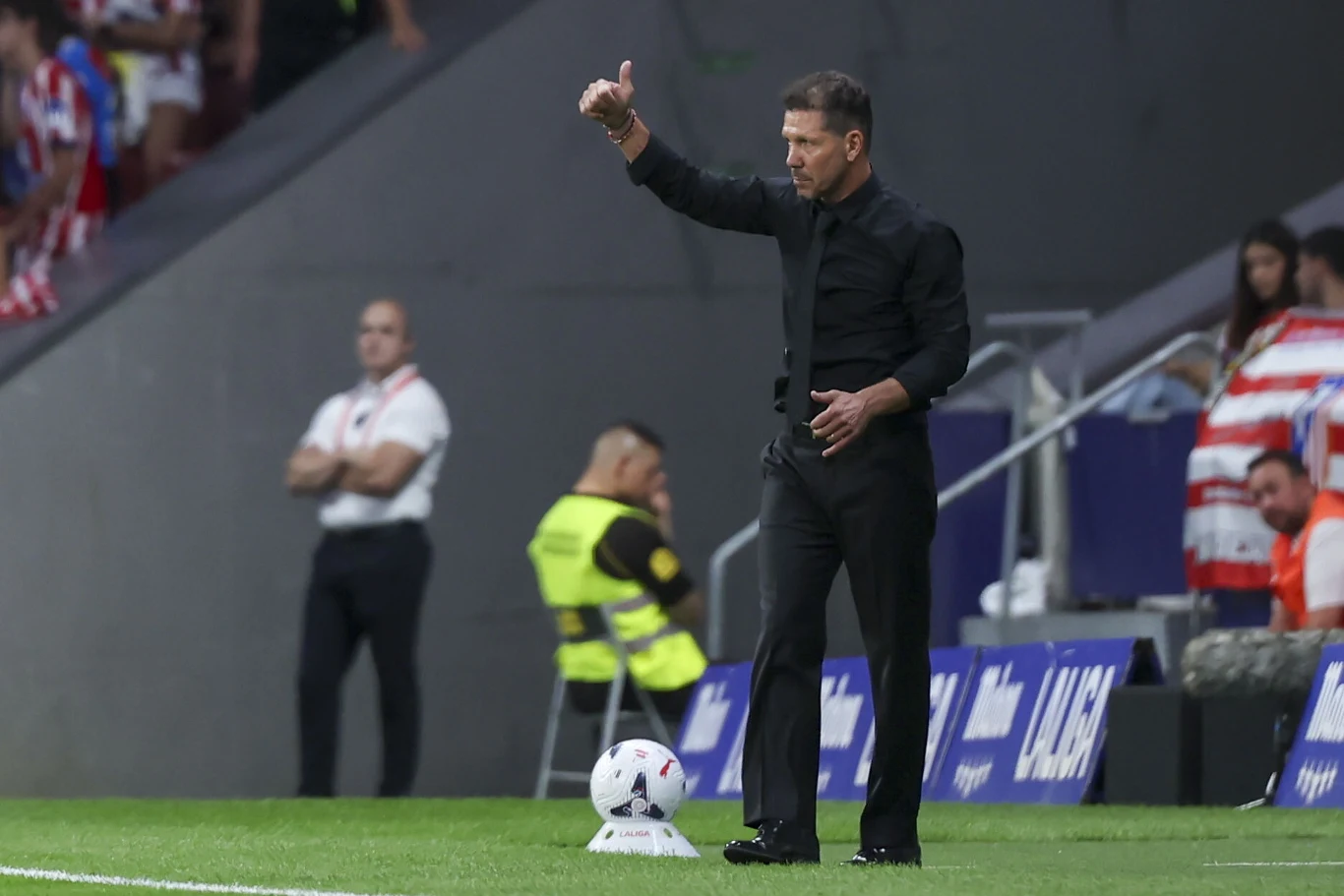 Trener Diego Simeone Mężczyzna w czarnym garniturze gestykuluje podnosząc kciuk do góry przy linii bocznej boiska piłkarskiego, obok leży piłka, w tle widoczni są stewardzi oraz fragment trybun z kibicami.