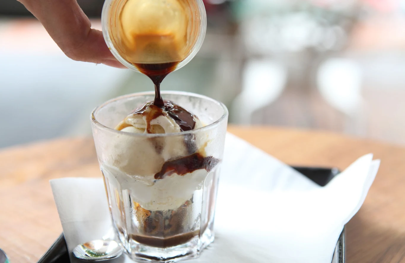 Affogato to włoski deser kawowy z dodatkiem gelato Affogato – porcja lodów waniliowych w szklance, polana espresso, na czarnej tacy z łyżeczką obok.