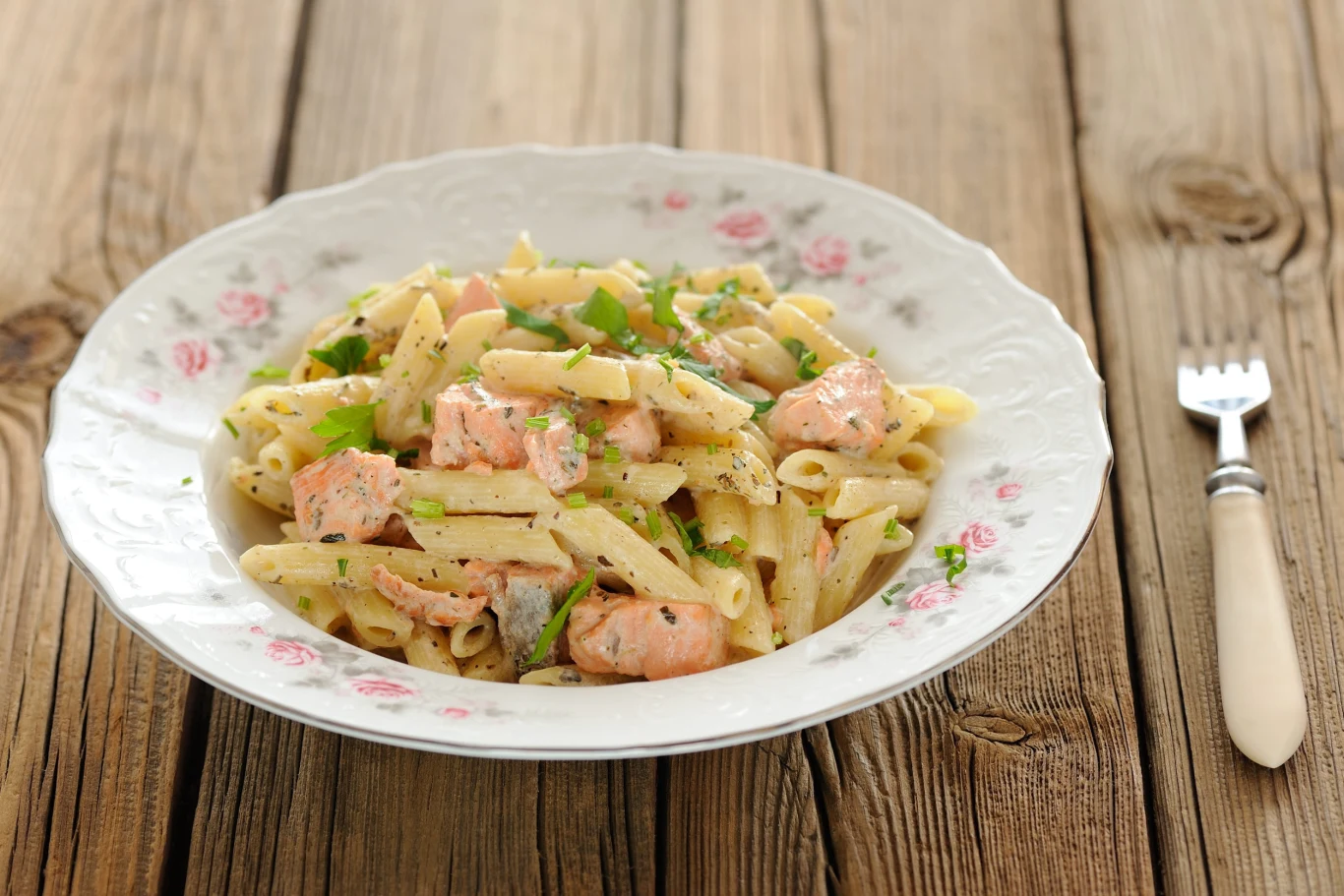Makaron z łososiem i cukinią to pożywny obiad do zrobienia w 20 minut Makaron penne z łososiem i cukinią, posypany pietruszką, w kremowym sosie na talerzu na drewnianym stole.