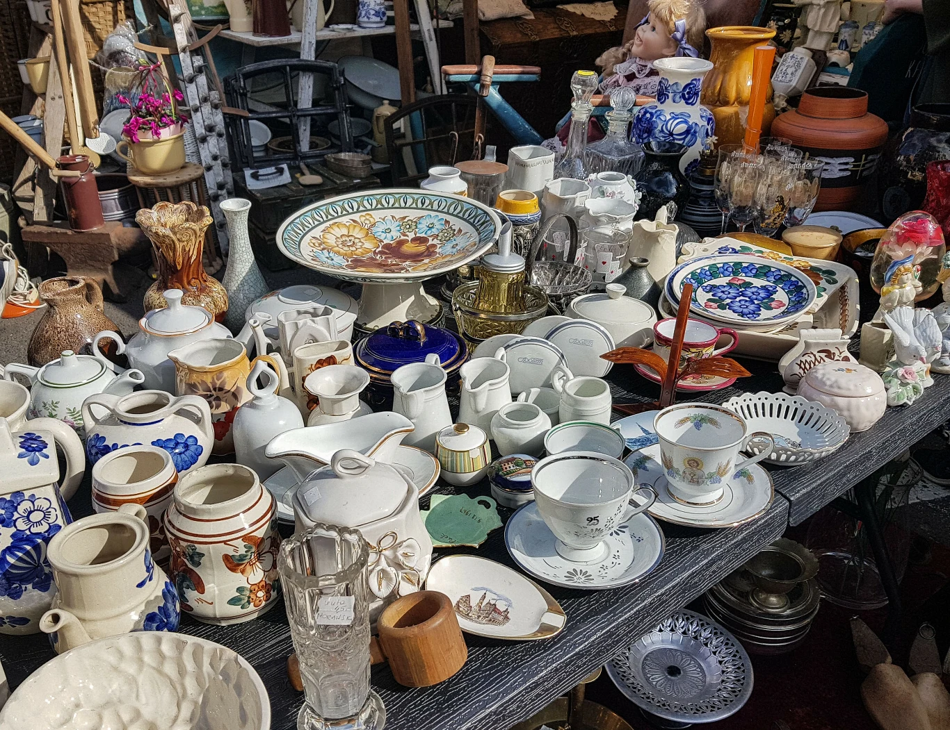 Stoisko na pchlim targu zastawione różnorodną porcelaną i ceramiką, ozdobnymi talerzami, dzbankami, filiżankami, szklankami i kieliszkami, a także kolorowymi wazonami oraz figurkami.