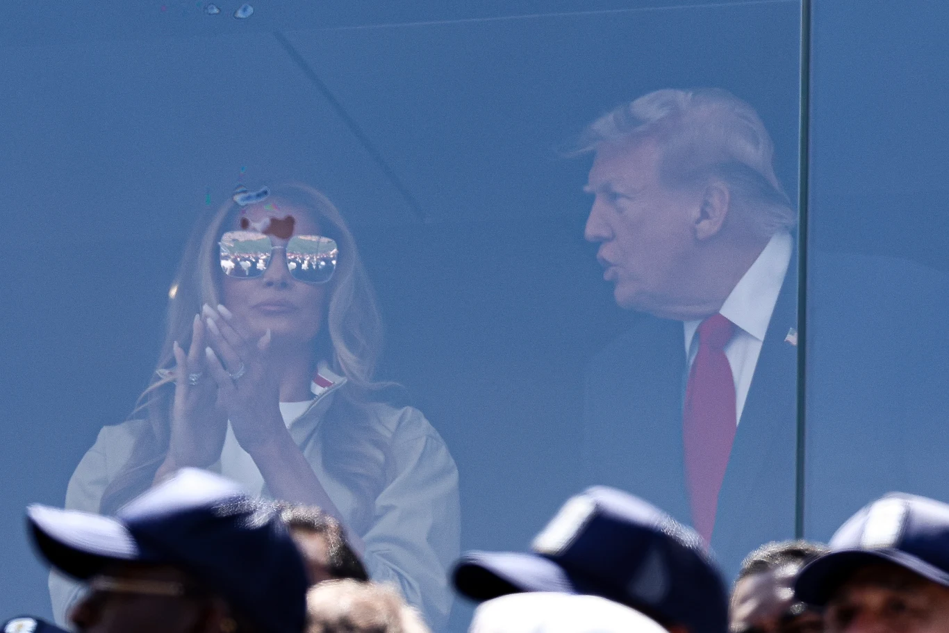 Melania Trump ostatnio była widziana publicznie u boku męża podczas finału Mistrzostw Świata FIFA Kobieta w okularach przeciwsłonecznych klaszcze, obok niej mężczyzna w garniturze i czerwonym krawacie, oboje za szybą, z przodu widoczne sylwetki ludzi w czapkach.