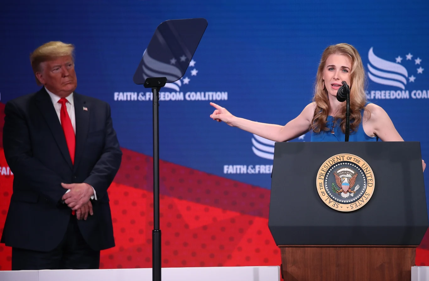 Natalie Harp wiernie stoi u boku Trumpa od dłuższego czasu Kobieta przemawia przy mównicy z prezydenckim emblematem, gestykulując ręką, podczas gdy mężczyzna w garniturze obserwuje ją z boku na tle niebiesko-czerwonej sceny z logotypem Faith & Freedom Coalition.