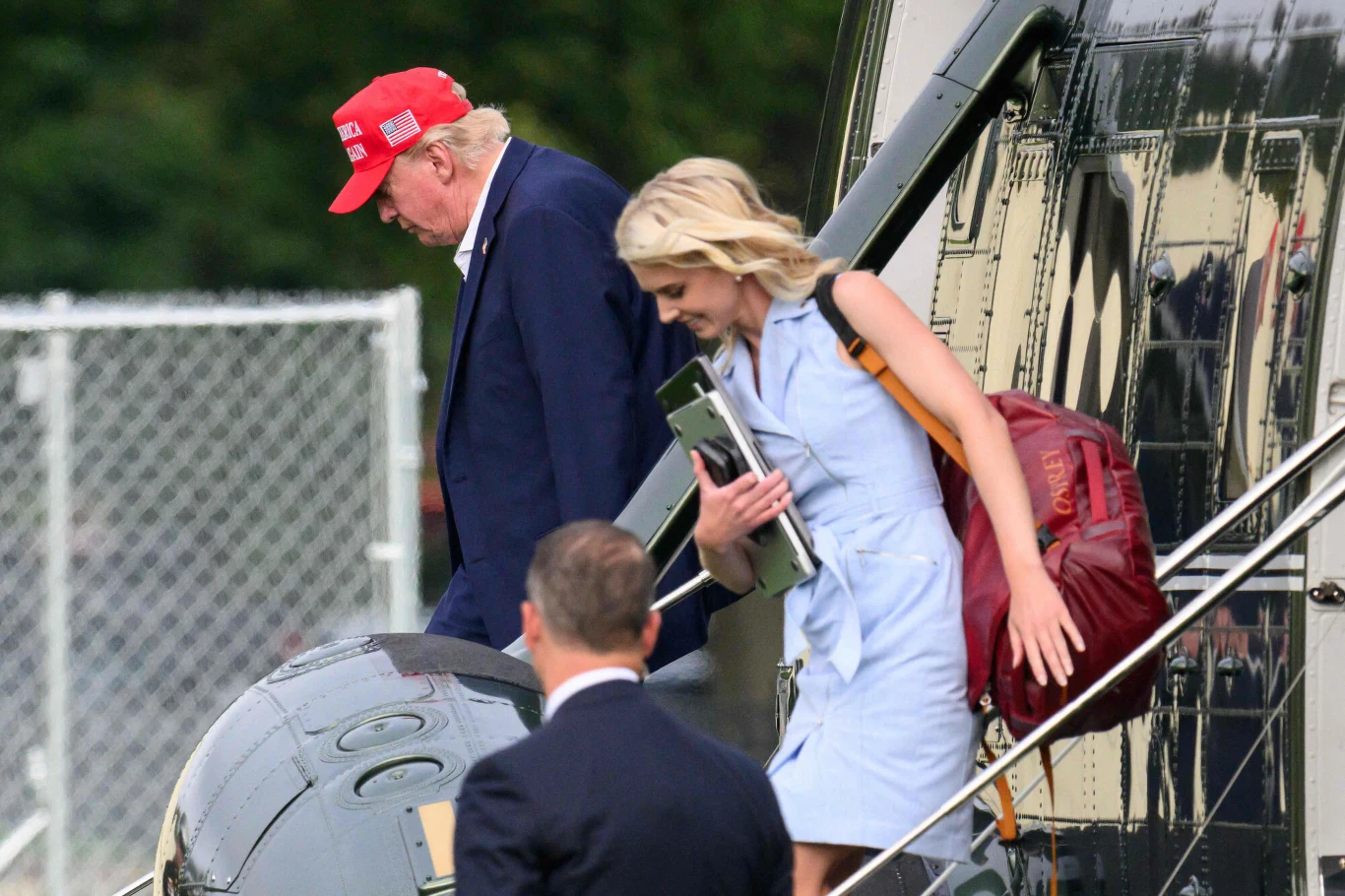 To nie Melania ostatnio jest widywana z Trumpem, a Natalie Harp, która nie opuszcza go nawet o krok Mężczyzna w czerwonej czapce schodzi po schodach śmigłowca, za nim kobieta w jasnej sukience z czerwonym plecakiem i dokumentami w ręku, przed wejściem do samolotu stoi mężczyzna w garniturze.