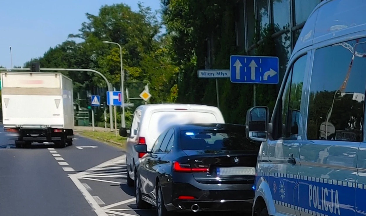 Radiowóz policyjny oraz dwa pojazdy osobowe zatrzymane na pasie przy jednym z głównych skrzyżowań, w tle widoczny znak drogowy kierujący ruch na dwa pasy.