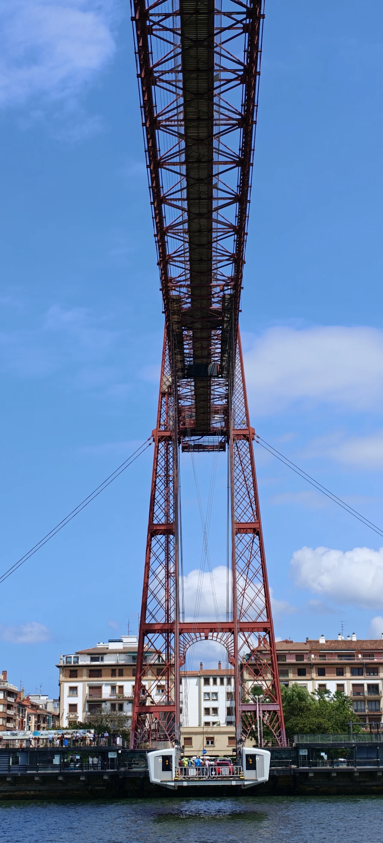 Czerwony stalowy most wiszący nad rzeką, a pod nim gondola przewożąca ludzi między dwoma brzegami wśród miejskiej zabudowy.