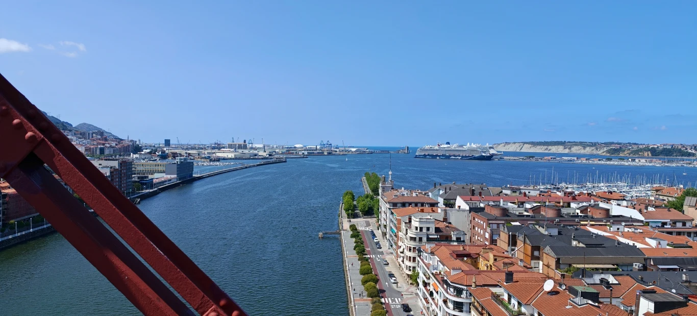 Panorama rzeki wpadającej do zatoki, z widocznymi zabudowaniami miejskimi po prawej stronie, fragmentem konstrukcji mostu po lewej oraz portem i cumującymi statkami w tle.
