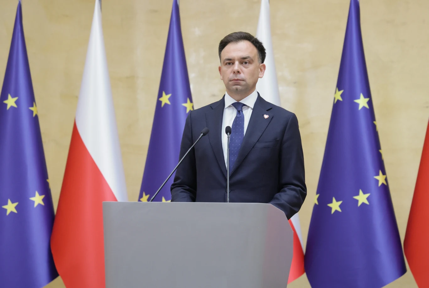 Minister finansów Andrzej Domański w trakcie konferencji prasowej dot. zmian w podatkach Minister Andrzej Domański przemawia przy mównicy na tle flag Polski i Unii Europejskiej podczas konferencji prasowej.