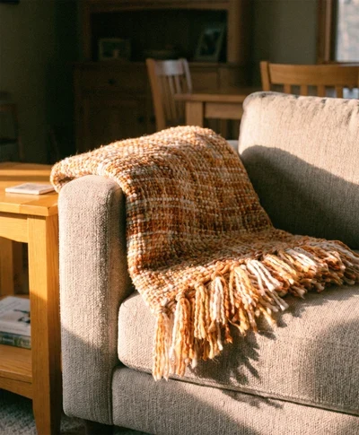 Poduszka dekoracyjna ruda gruby prążek za 20 zł Beżowa sofa z brązowym, plecionym kocem o długich frędzlach, ustawiona obok drewnianego stolika w jasnym, przytulnym pomieszczeniu.