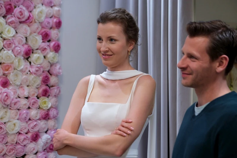 Adam będzie zachwycony widokiem Natalii w ślubnej sukni Uśmiechnięta kobieta w eleganckiej białej sukni stoi obok mężczyzny, na tle ściany z jasnoróżowymi i białymi kwiatami.
