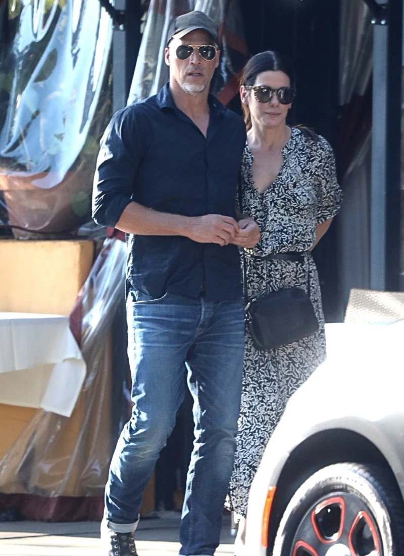 Sandra Bullock o śmierci Bryana Randalla. "Nie wolno mi było o tym mówić" Para dorosłych stoi razem na chodniku, mężczyzna ubrany w ciemną koszulę, spodnie jeansowe, czapkę z daszkiem i okulary przeciwsłoneczne, kobieta w sukience z czarno-białym wzorem, okularach przeciwsłonecznych i z małą czarną torebką na ramieniu; w kad...