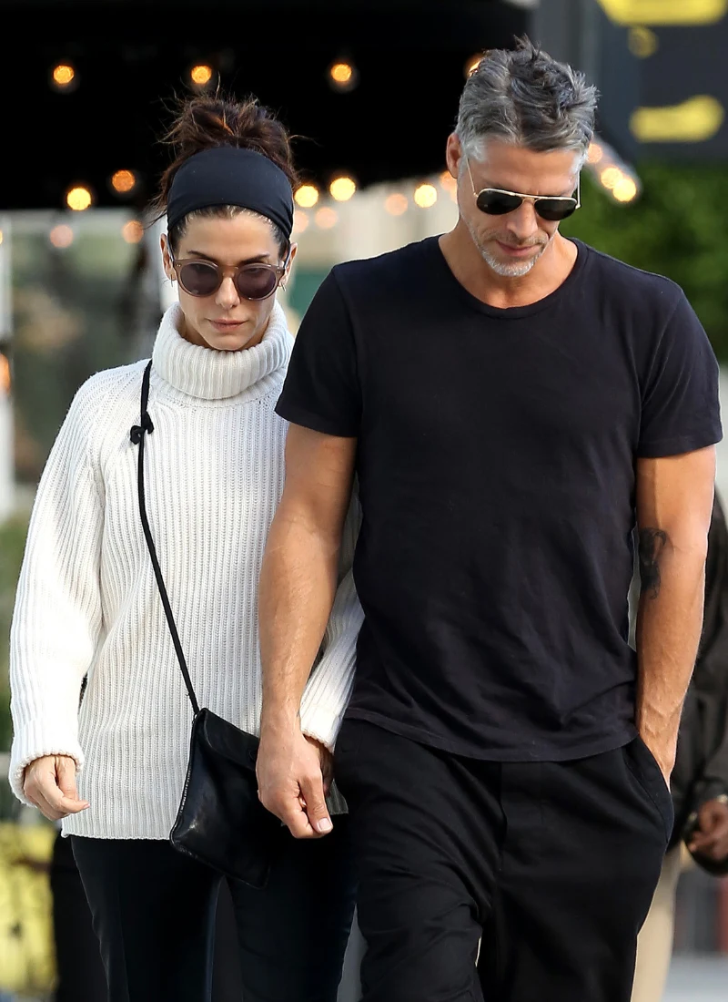 Sandra Bullock i Bryan Randall Para dorosłych w ciemnych okularach spaceruje razem, kobieta ma na sobie biały sweter i czarną torebkę, mężczyzna ubrany jest na czarno.