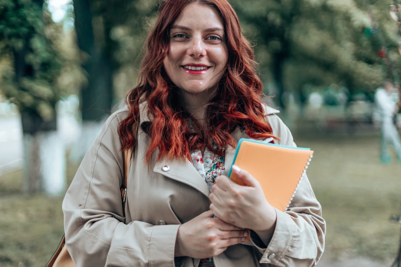 Wrzesień otworzy przed nimi drzwi do sukcesu. Te trzy znaki zodiaku mają szansę na przełom Młoda kobieta z rudymi włosami w beżowym płaszczu, trzymająca zeszyty i książki, stoi w zielonym parku.