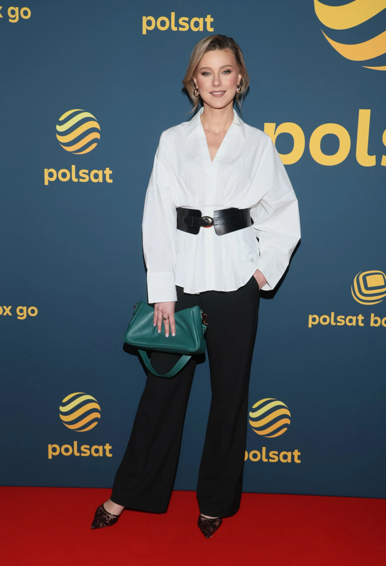 Szeroki pasek doskonale prezentuje się z koszulą bądź swetrem Kobieta w białej koszuli z szerokim czarnym paskiem, czarnych szerokich spodniach i eleganckich butach, trzymająca zieloną torebkę, pozuje na czerwonym dywanie na tle z logo Polsatu.