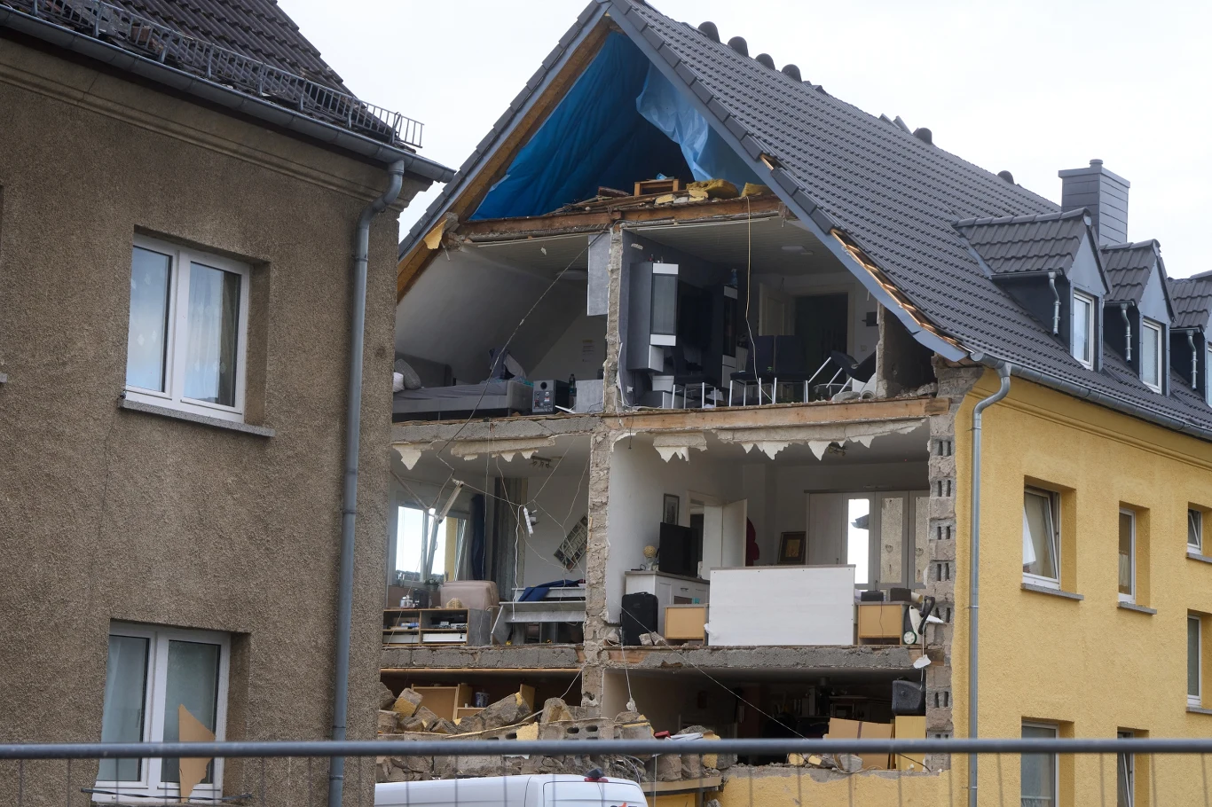 Zniszczona kamienica z odsłoniętymi kondygnacjami, zawalonym stropem i rozrzuconymi meblami wewnątrz, ślady katastrofy budowlanej obok stojących nienaruszonych budynków.