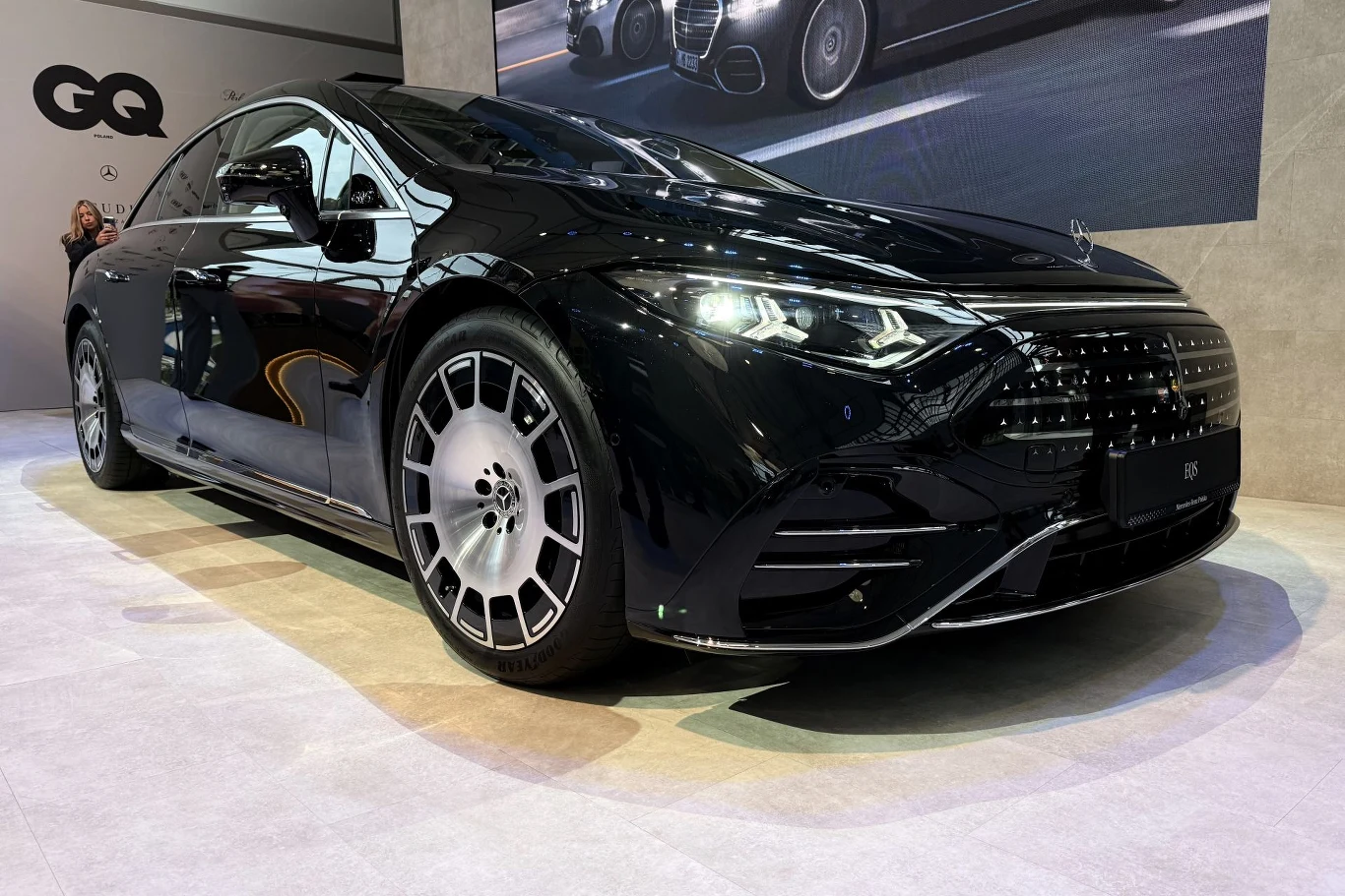 Mercedes EQS opcjonalnie będzie wyposażony w system steer-by-wire. W takiej specyfikacji kierować będziemy wolantem. W ofercie wciąż będzie jednak elektromechaniczny układ kierowniczy z normalną kierownicą. Czarny luksusowy sedan o nowoczesnym, aerodynamicznym kształcie nadwozia, charakterystycznych dużych felgach i eleganckim wykończeniu karoserii, ustawiony w jasnym salonie wystawowym.