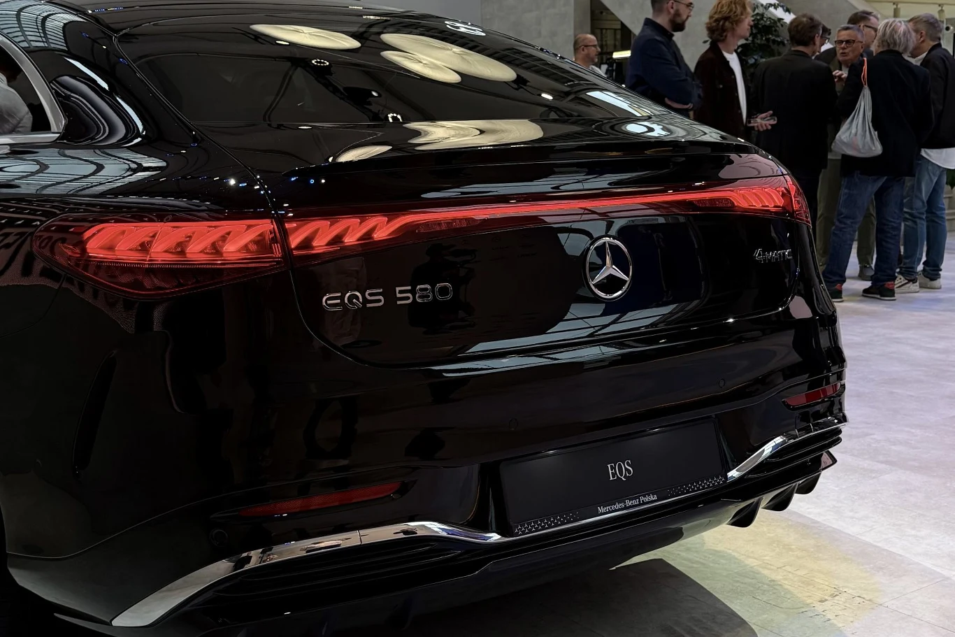 Mercedes EQS dostępny w trzech wariantach napędu: od 408 do 585 KM. Na jednym ładowaniu ma przejechać nawet 919 km. Czarny Mercedes EQS 580 4MATIC z charakterystyczną linią tylnych świateł LED, otoczony grupą osób podczas prezentacji w jasno oświetlonym wnętrzu.