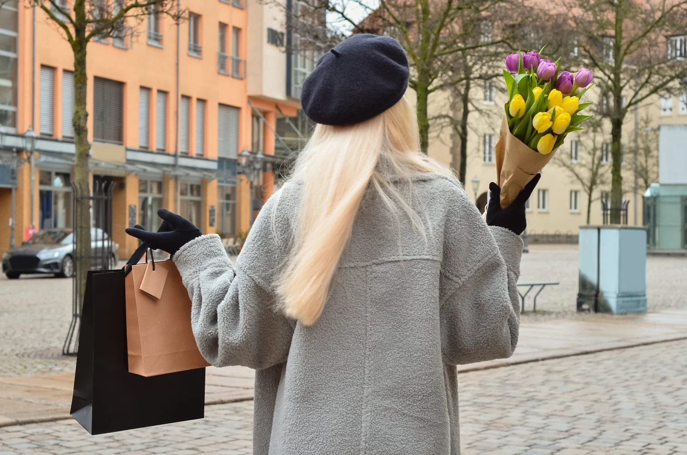 Ostatnio furorę w mediach społecznościowych robi trend "kinda chic" Kobieta z długimi blond włosami w szarym płaszczu i czarnym berecie trzyma bukiet tulipanów i torby zakupowe na miejskim rynku.