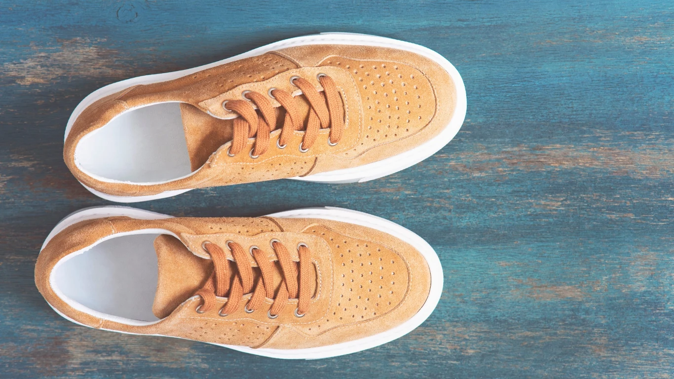 Buty sportowe w eleganckim wydani podkreślą jesienny look Para jasnobrązowych zamszowych sneakersów z białą podeszwą ustawiona na niebieskiej drewnianej powierzchni.