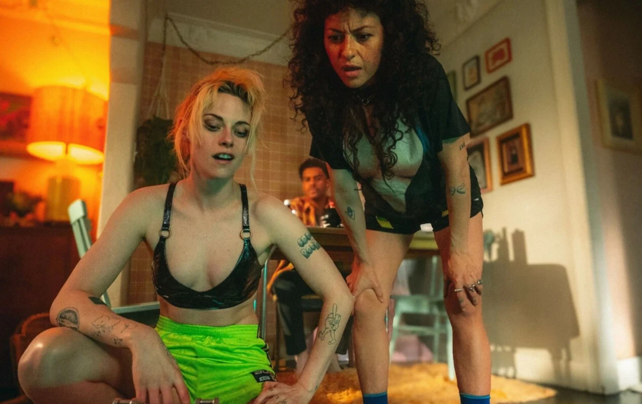 Kadr z filmu "Wrong Girls" Dwie młode kobiety w sportowych ubraniach pochylone nad czymś poza kadrem, w tle mężczyzna przy stole.