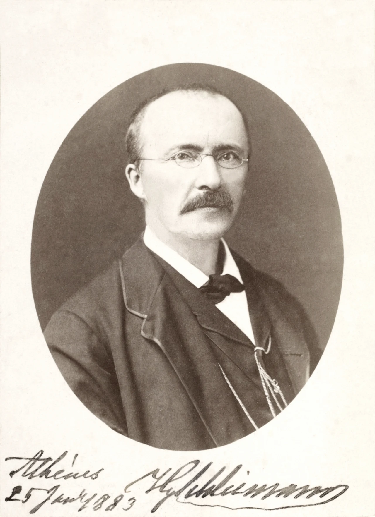 Portret Heinricha Schliemanna z 1883 roku Mężczyzna w średnim wieku z wąsami i okularami, ubrany w marynarkę, na białym tle z owalną ramką i podpisem pod zdjęciem.